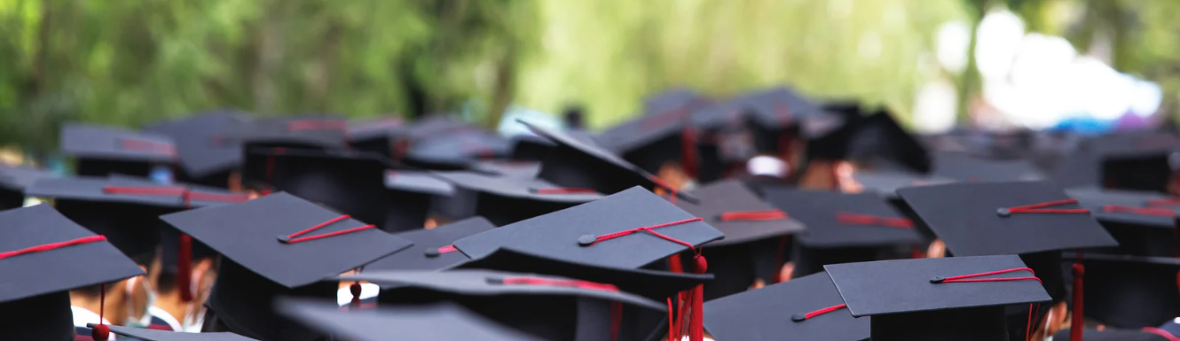 Photo of black graduation caps with red tassels in front of green trees blurred in the background.