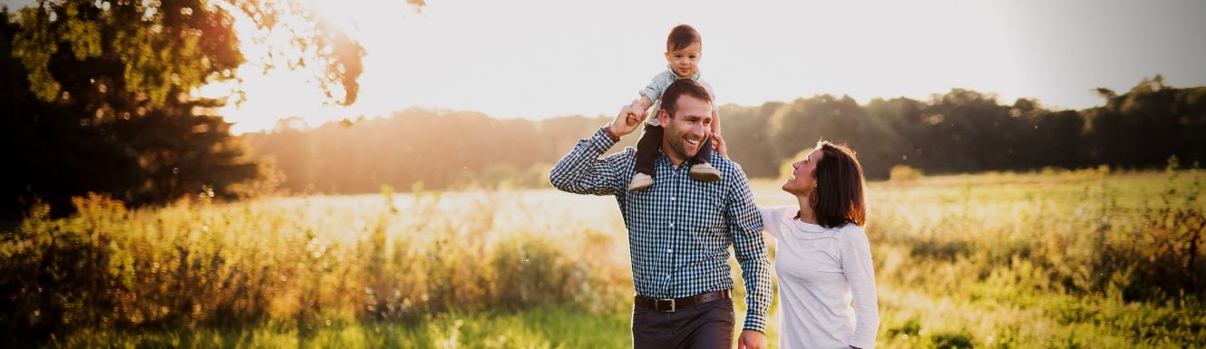 Man walking in field with a baby on his shoulders and holding on to woman's hand. Woman is smiling up at the baby.